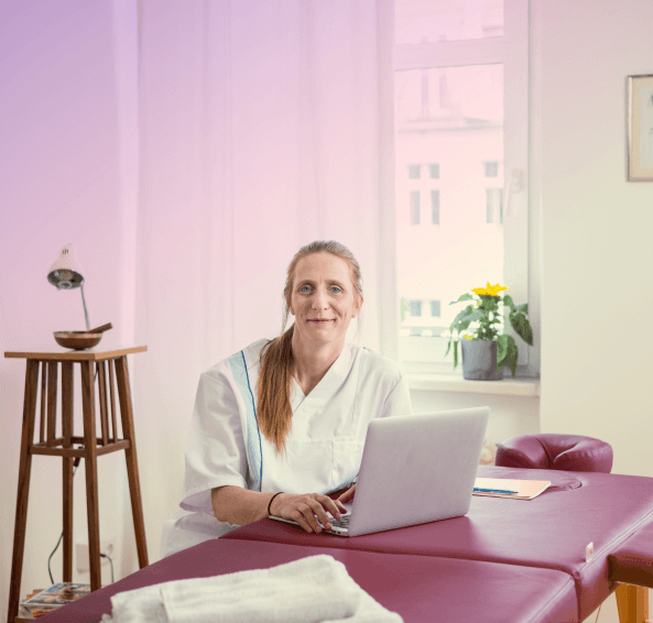 Eine Physiotherapeutin erledigt ihre Büroarbeit auf der Massageliege und schaut in die Kamera.